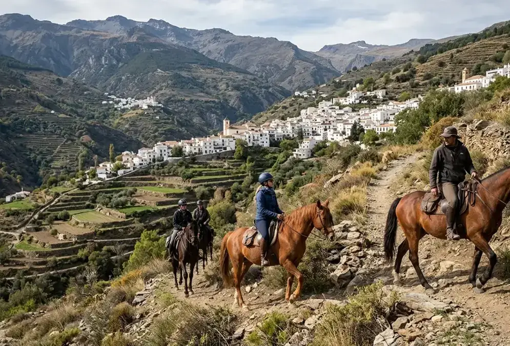 Riders on horseback on a mountain trail in the Alpujarras with white village rooftops and Sierra Nevada peaks in the background
