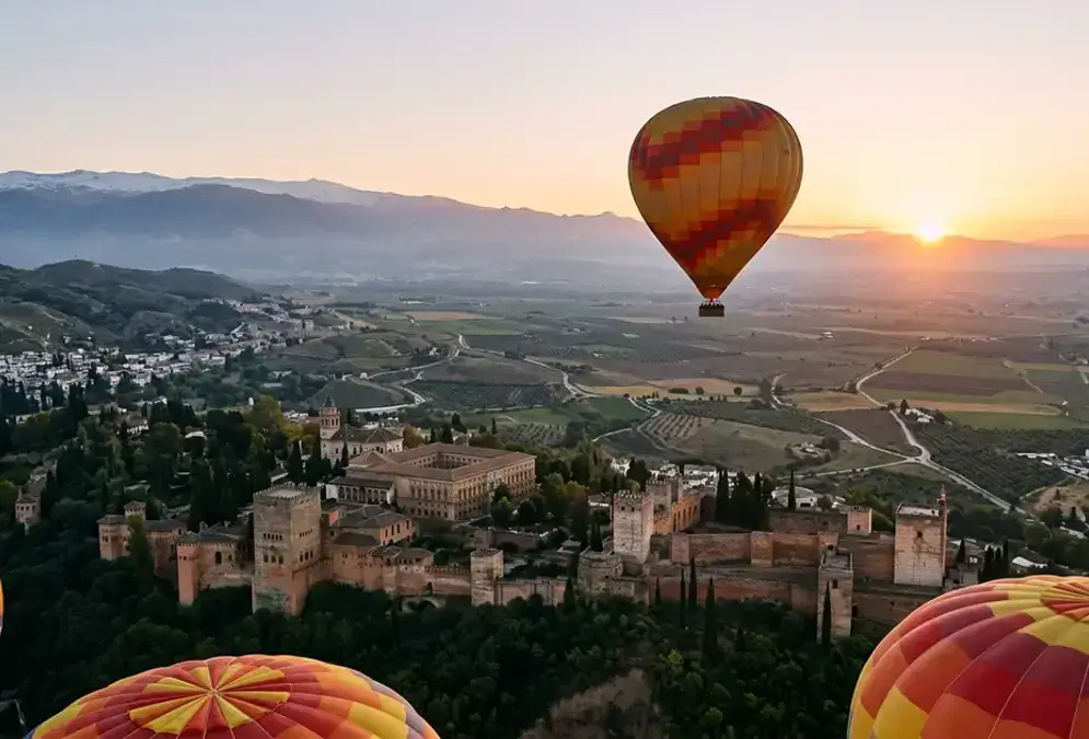 Hot air balloon floating above the Vega de Granada at sunrise, Alhambra on its hilltop visible in the middle distance with the Sierra Nevada peaks behind