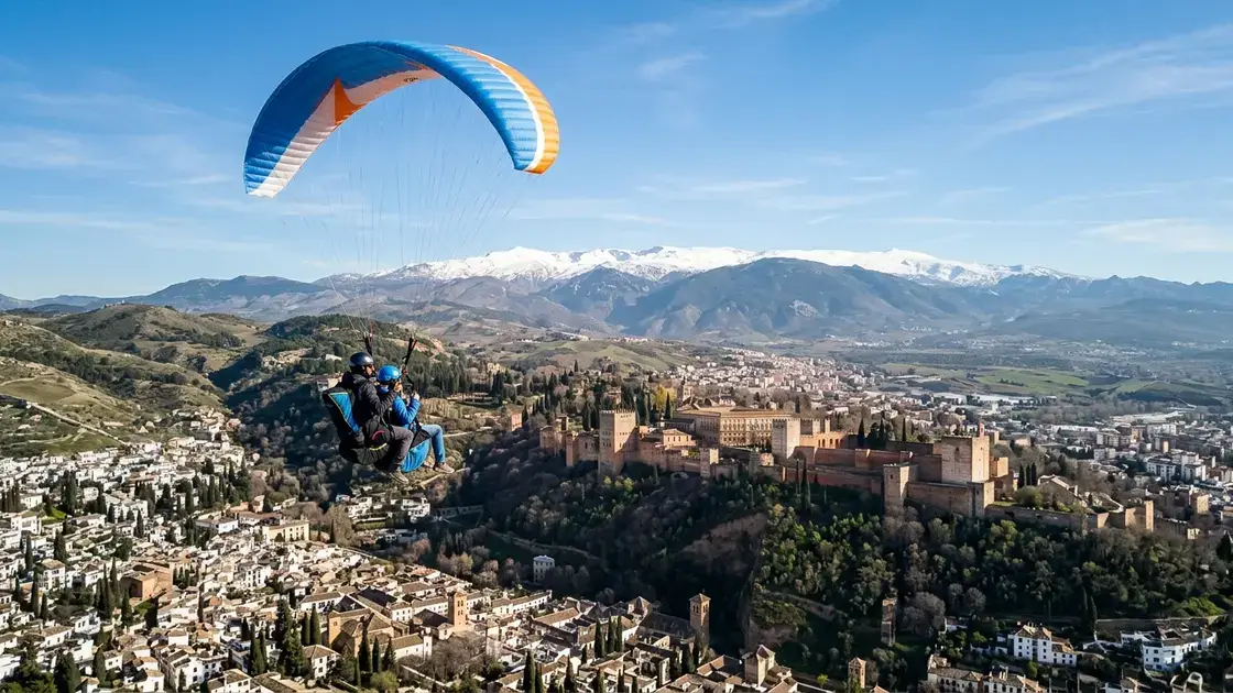 Paraglider soaring above Granada's Sierra Nevada mountains with snow-capped peaks, the Alhambra palace, and Cenes de la Vega valley visible below in morning light