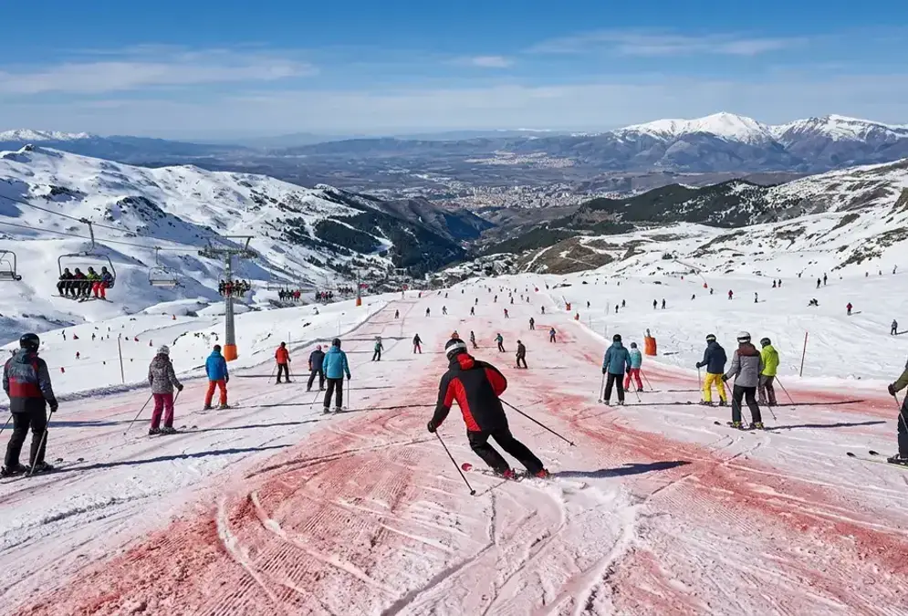 Skiers on a wide red piste at Sierra Nevada ski resort with Granada plain visible far below under blue sky